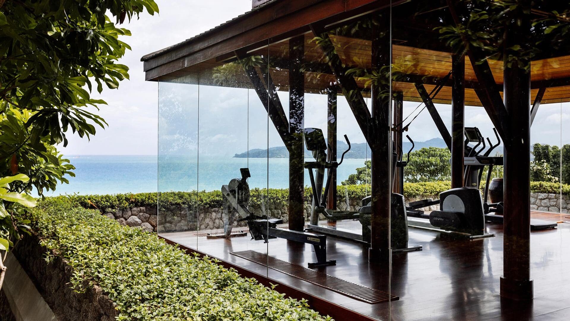 A glass-walled gym pavilion at Amanpuri in Thailand, set among lush greenery and overlooking the ocean, with cardio machines and weight equipment arranged on dark wood floors beneath a timber-framed roof, blending indoor training with open coastal views.
