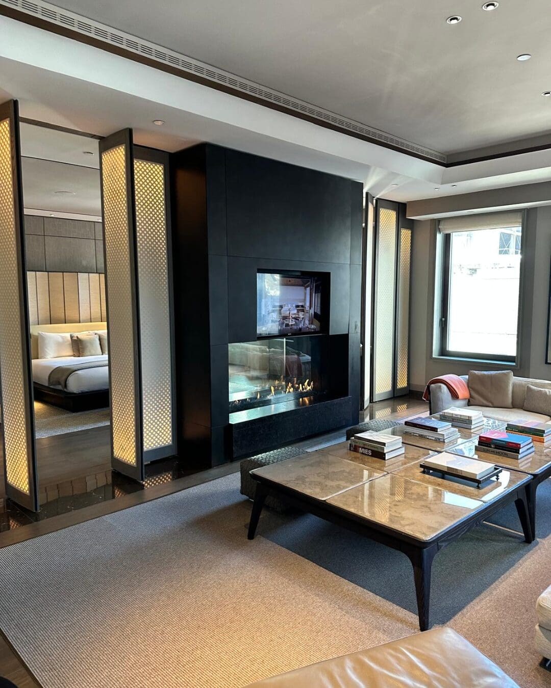 Aman New York suite living room with fireplace, neutral furnishings, and city views through floor-to-ceiling windows