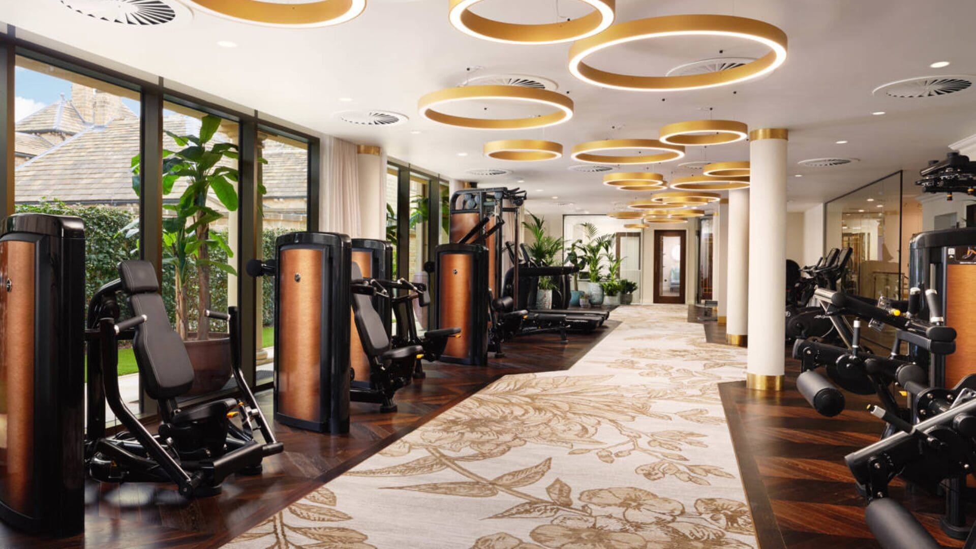 Elegant gym at Grantley Hall, Yorkshire featuring premium strength machines, large windows with garden views, and warm lighting with gold circular ceiling fixtures.