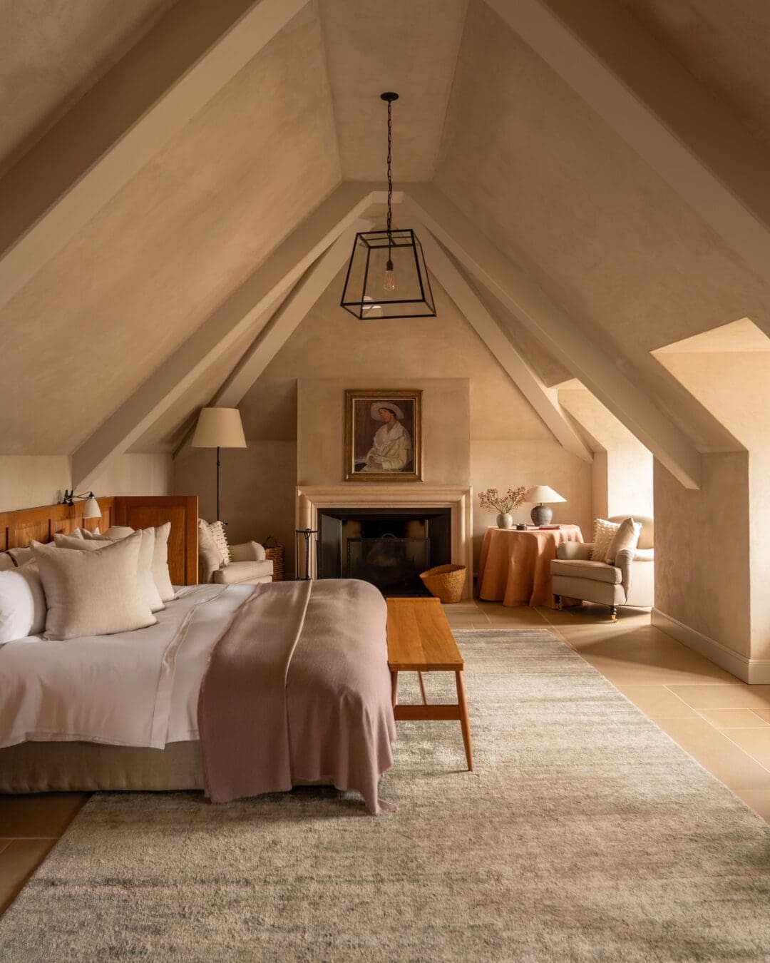 A calm, neutral-toned bedroom at Heckfield Place with vaulted ceilings, soft natural light, and understated furnishings centred around a fireplace.