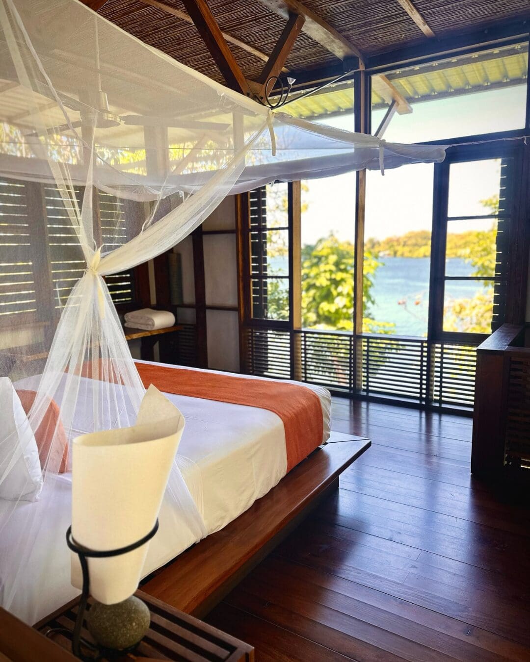 An open-air bedroom at Jicaro Private Island with wooden interiors, a canopy bed, and views out across the lake through large windows.