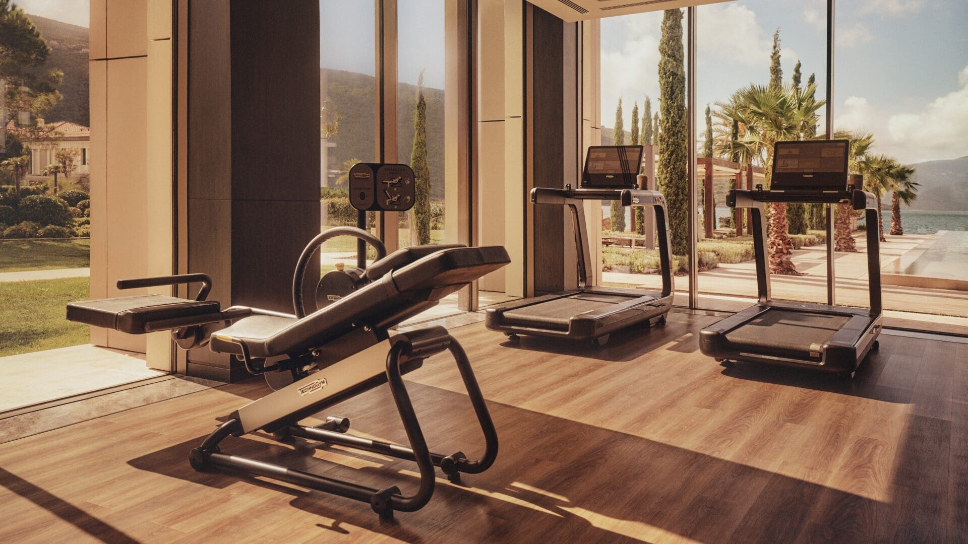 A sunlit, modern hotel gym at One&Only Portonovi in Montenegro, featuring treadmills and a weight bench set on warm wood flooring, with floor-to-ceiling windows opening onto palm trees, manicured gardens, and views of the Adriatic coast beyond.