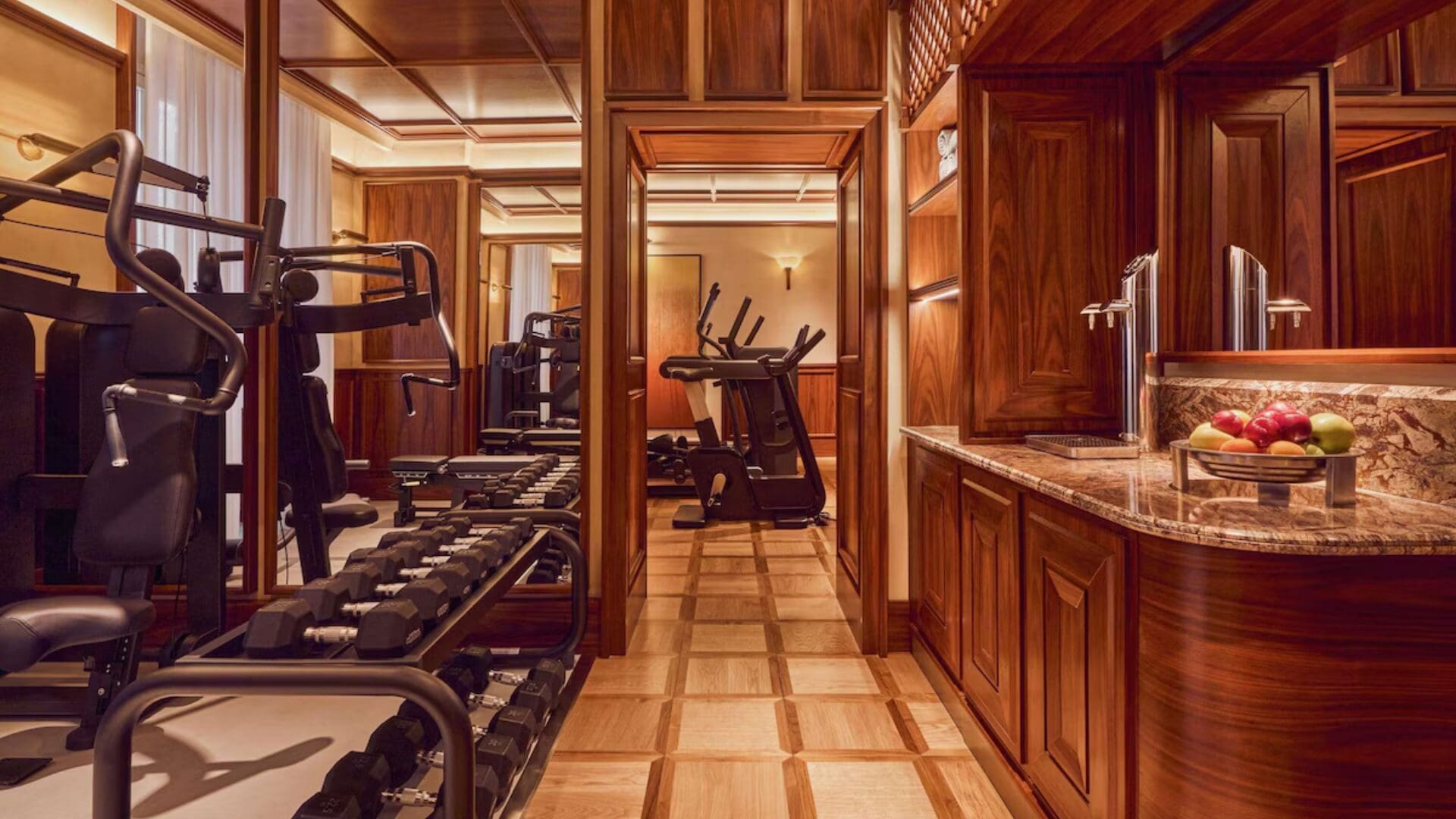 A warm, wood-panelled gym interior with classic detailing, featuring a row of dumbbells and weight machines on the left, cardio equipment visible through a doorway ahead, and a marble-topped counter with fresh fruit on the right, all set on a patterned parquet floor under soft ambient lighting.