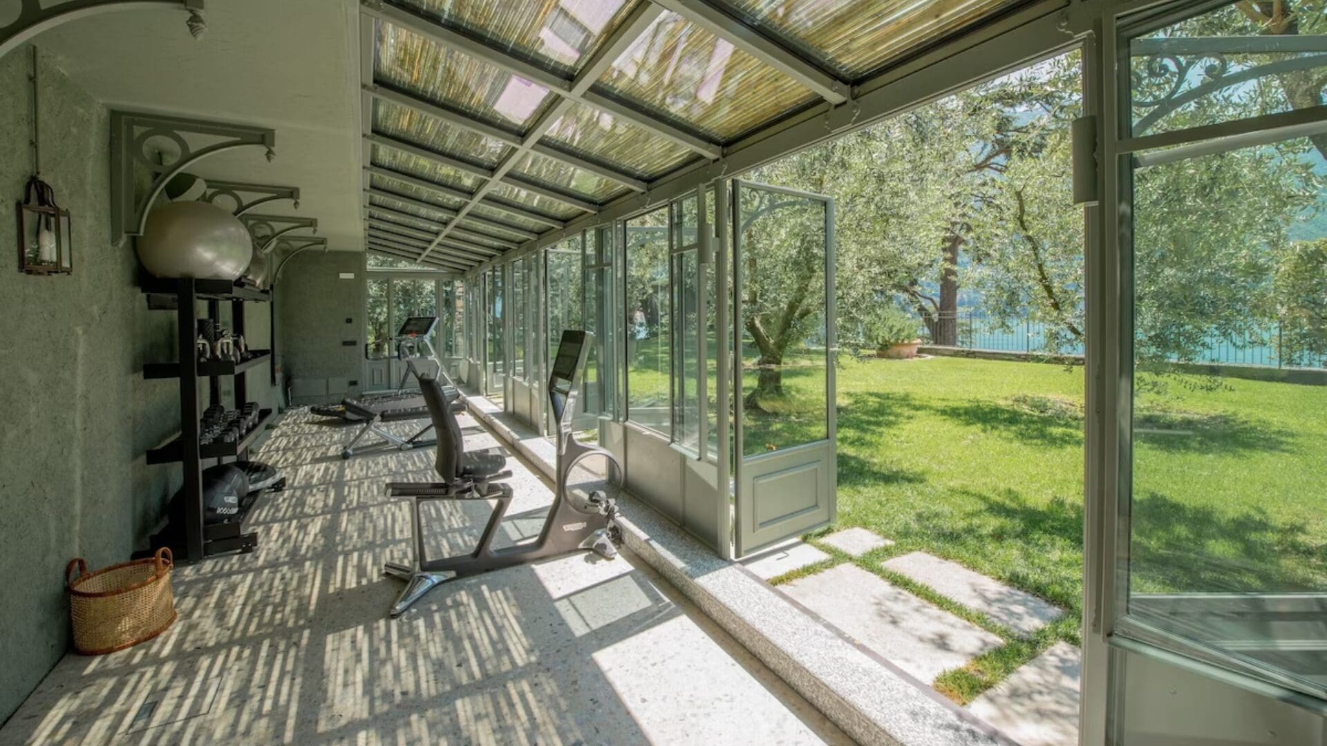Light-filled gym at Passalacqua, Lake Como, set within a glass conservatory overlooking the gardens and lake, with simple equipment and a relaxed, open-air feel.
