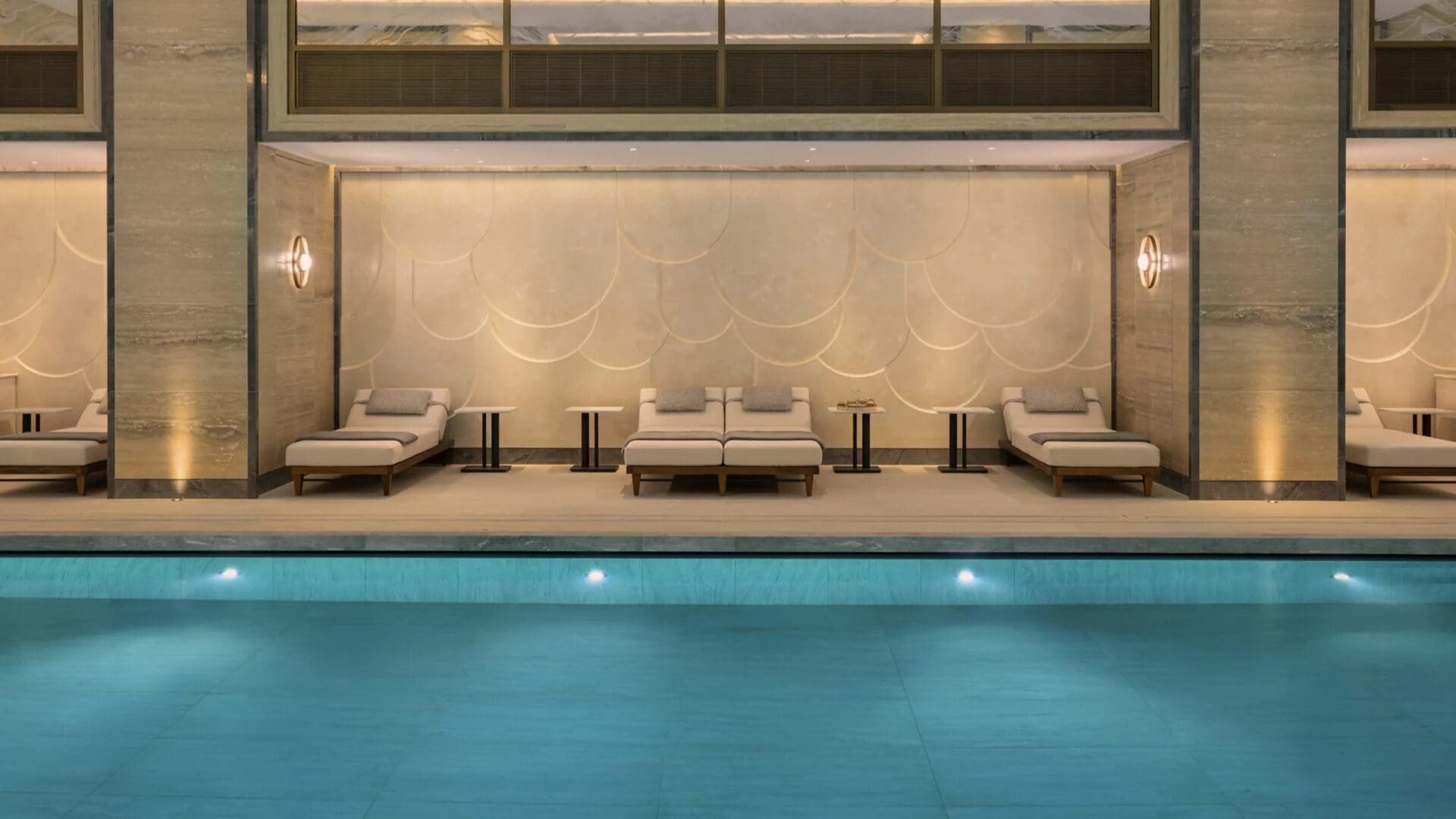 Indoor pool at Raffles London at The OWO, with calm blue water, marble walls, and a row of cushioned loungers set beneath soft, ambient lighting.