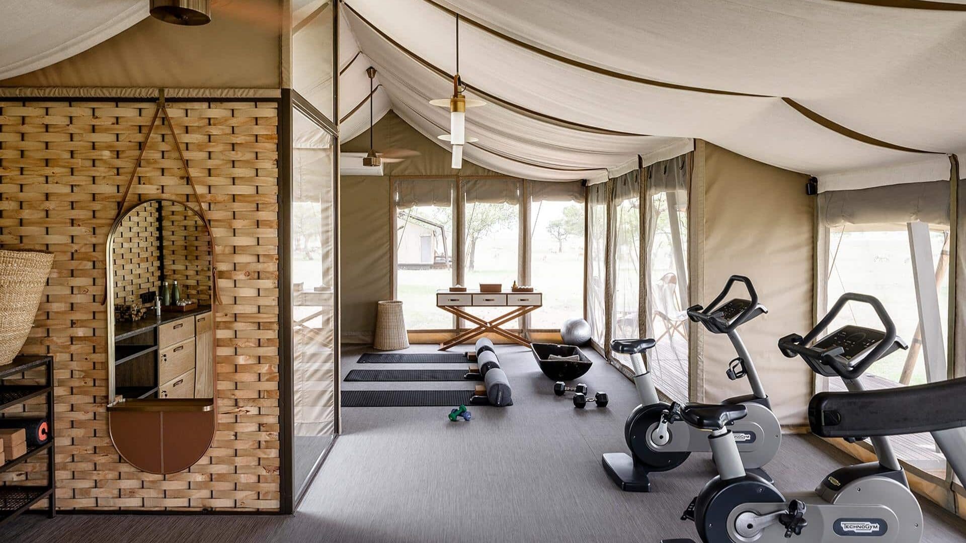 A light-filled canvas gym tent at Singita Sabora in Tanzania, featuring stationary bikes, yoga mats, and free weights arranged on a soft grey floor, with woven textures, warm timber accents, and open views across the Serengeti plains through wide mesh windows.