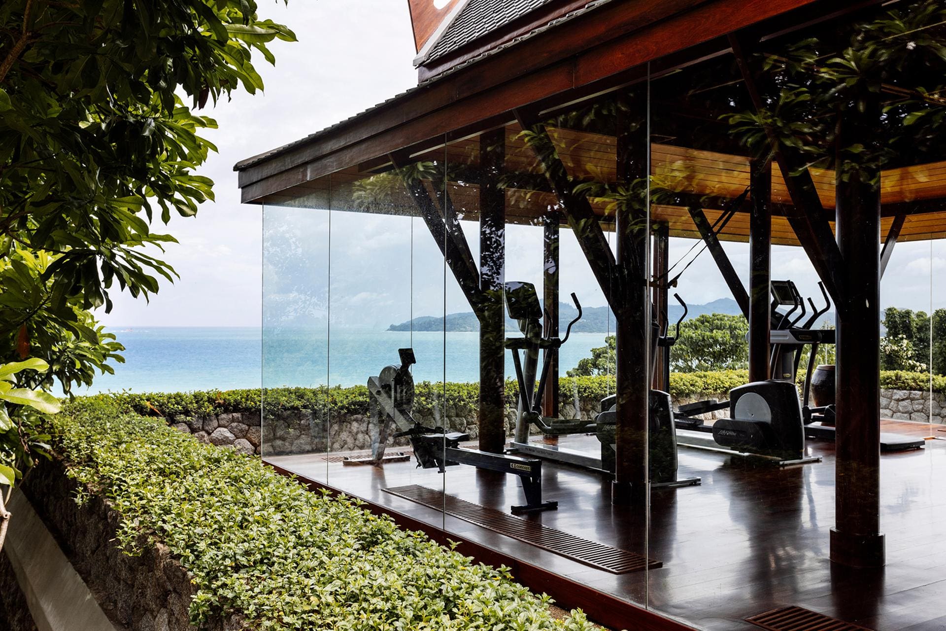 A glass-walled gym pavilion at Amanpuri in Thailand, set among lush greenery and overlooking the ocean, with cardio machines and weight equipment arranged on dark wood floors beneath a timber-framed roof, blending indoor training with open coastal views.